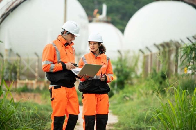 Deux ingénieurs en tenue orange et casque blanc consultent des documents pour trouver des chantiers sur site industriel