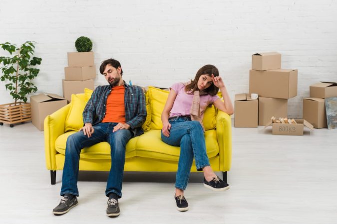 Un couple épuisé est assis sur un canapé jaune vif au milieu de cartons de déménagement, réfléchissant au choix d'un logement meublé ou nu.