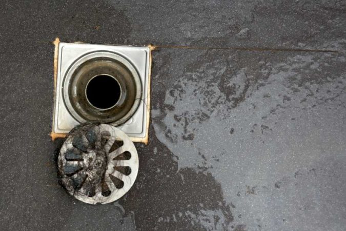 Gros plan sur un siphon de sol de douche carré en métal rouillé, dont la grille est retirée et obstruée par un amas de cheveux et de résidus sombres.