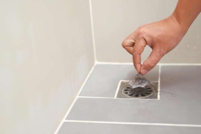 Une main retire délicatement un amas de cheveux et de résidus accumulés sur la grille d'un siphon de douche, une étape essentielle pour nettoyer les canalisations.